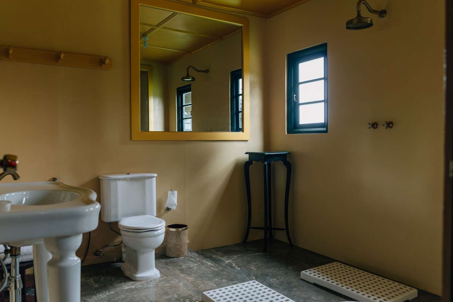 A plain cream bathroom with a pedestal basin, a gilt-framed mirror, a rain shower and duckboards on the floor.