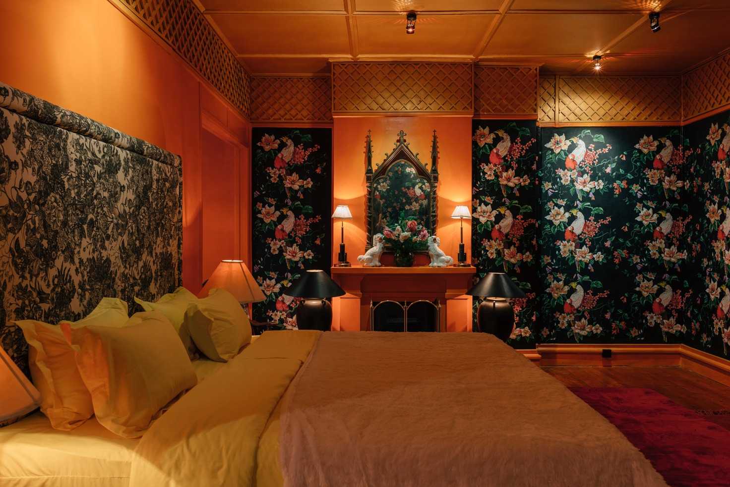 A saffron bedroom: a toile headboard, yellow linen, parrot wallpaper on deep green and a Gothic mirror over the mantel.