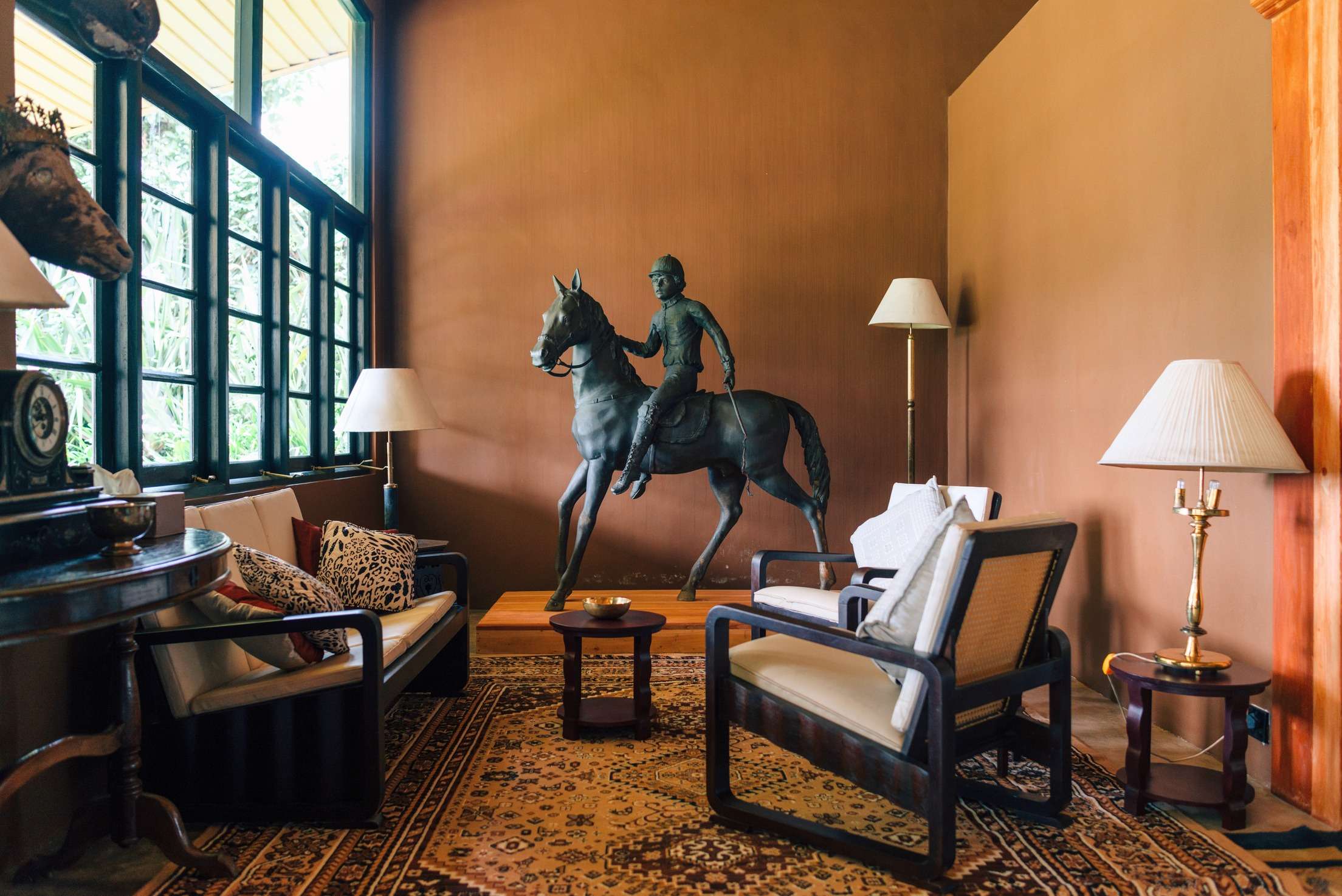 A warm brown sitting room with a large statue of a horse and jockey mid-stride, cane-backed armchairs and tall lamps.