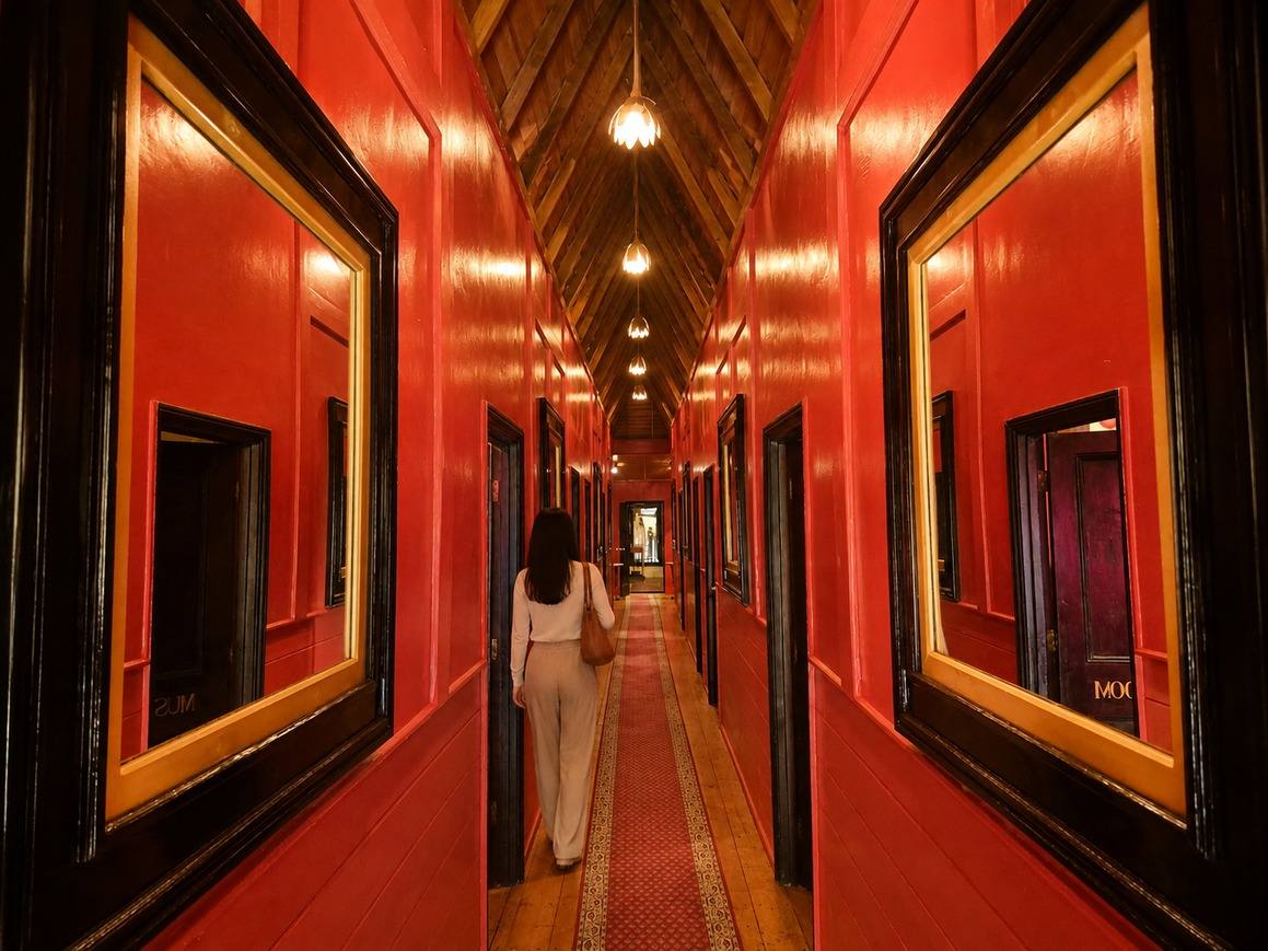 The long red corridor: lacquered walls, framed mirrors on both sides, a runner, and lamps hanging from a pitched timber ceiling.