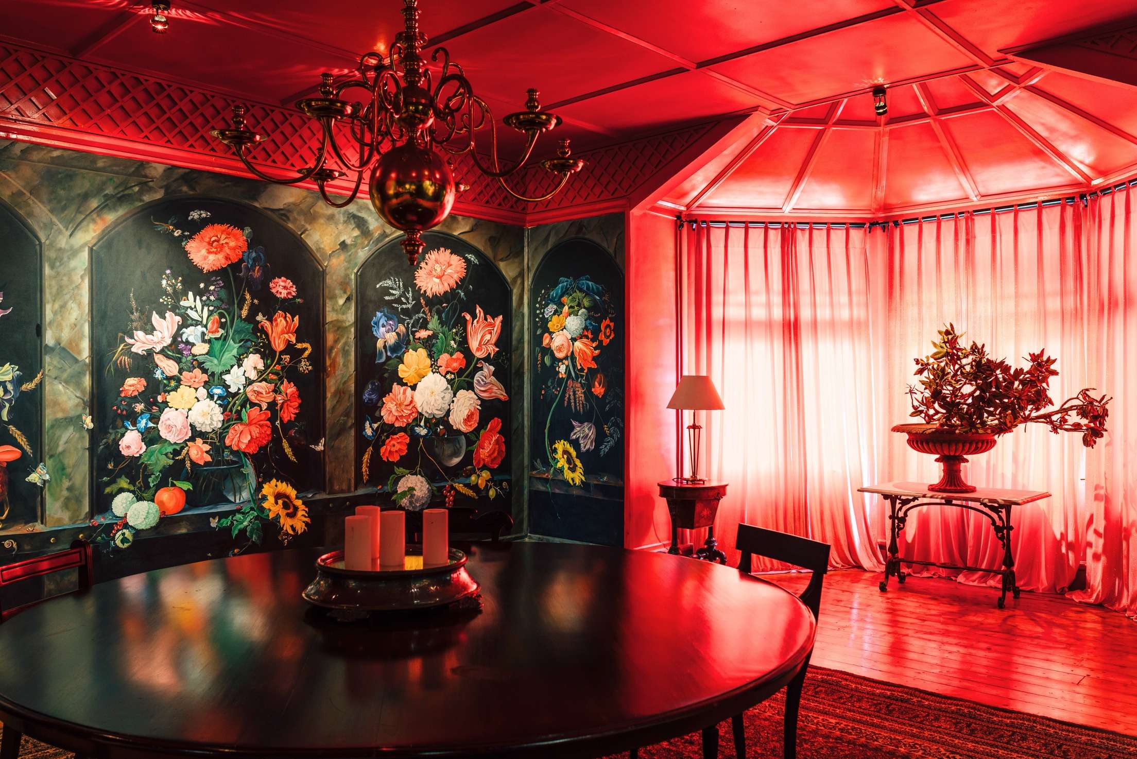 The red dining room: a round dark table, a brass chandelier, floral panels painted like Dutch still lifes and a bay window hung with red curtains.