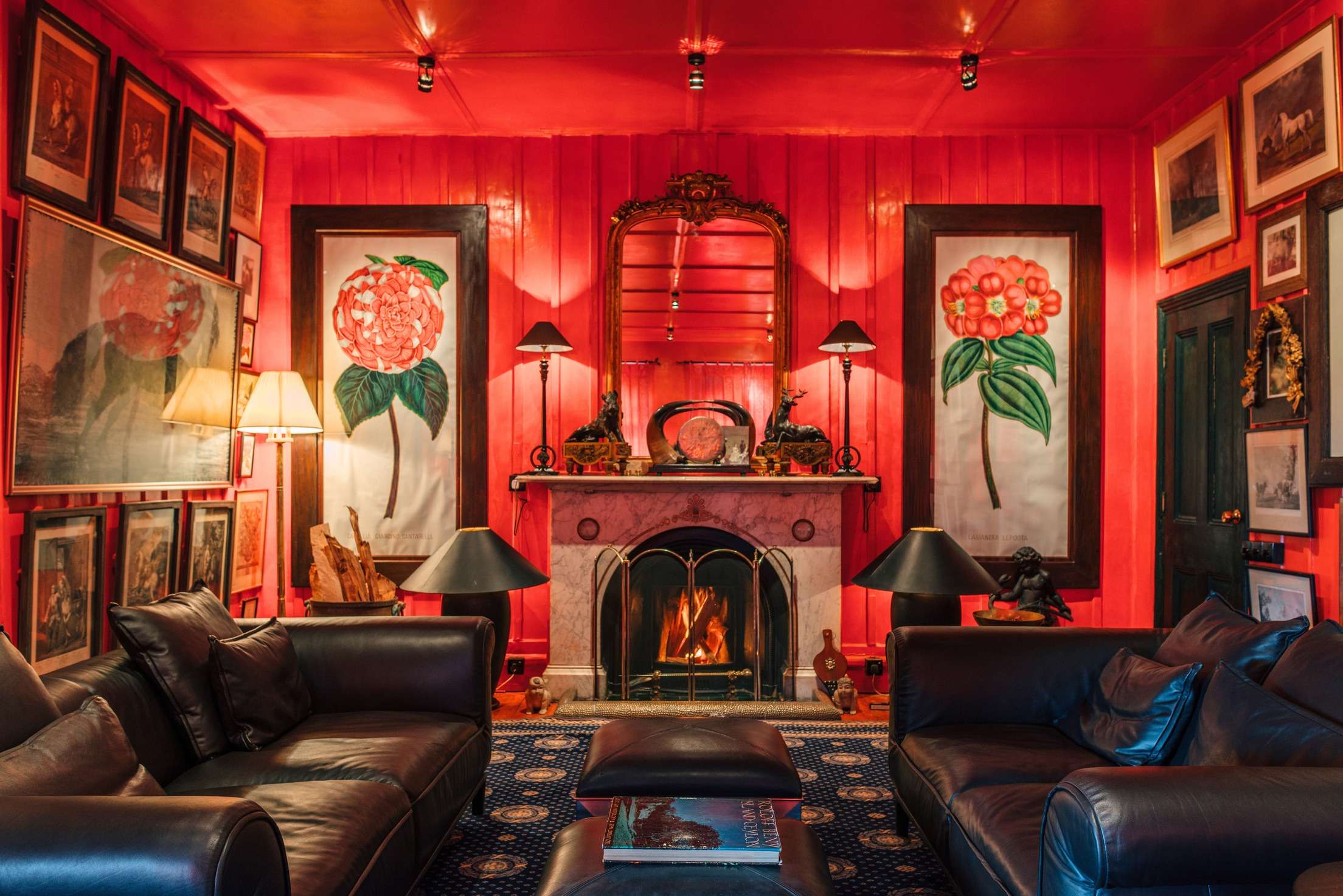The red drawing room: a marble fireplace with a fire lit, a gilt mirror above, botanical prints either side and leather sofas.
