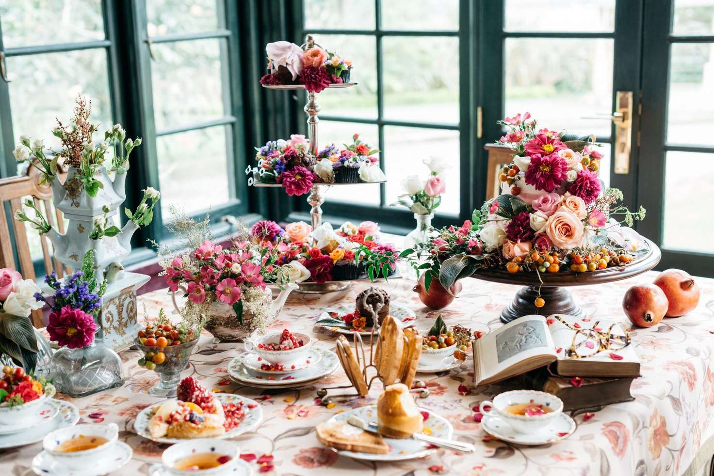 A high tea table crowded with flowers, cakes on tiered stands, teacups, pomegranates and an open book.