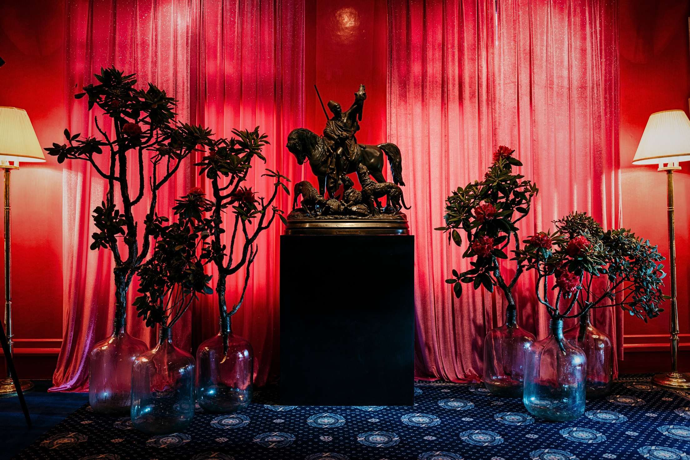 A dark horseman with hounds on a black plinth, before a red curtain, between lamps.