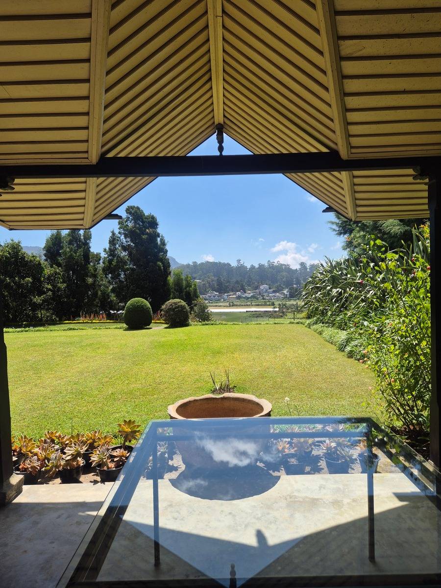 Looking out from the verandah: a wide green lawn, an urn and distant hills under a blue sky.