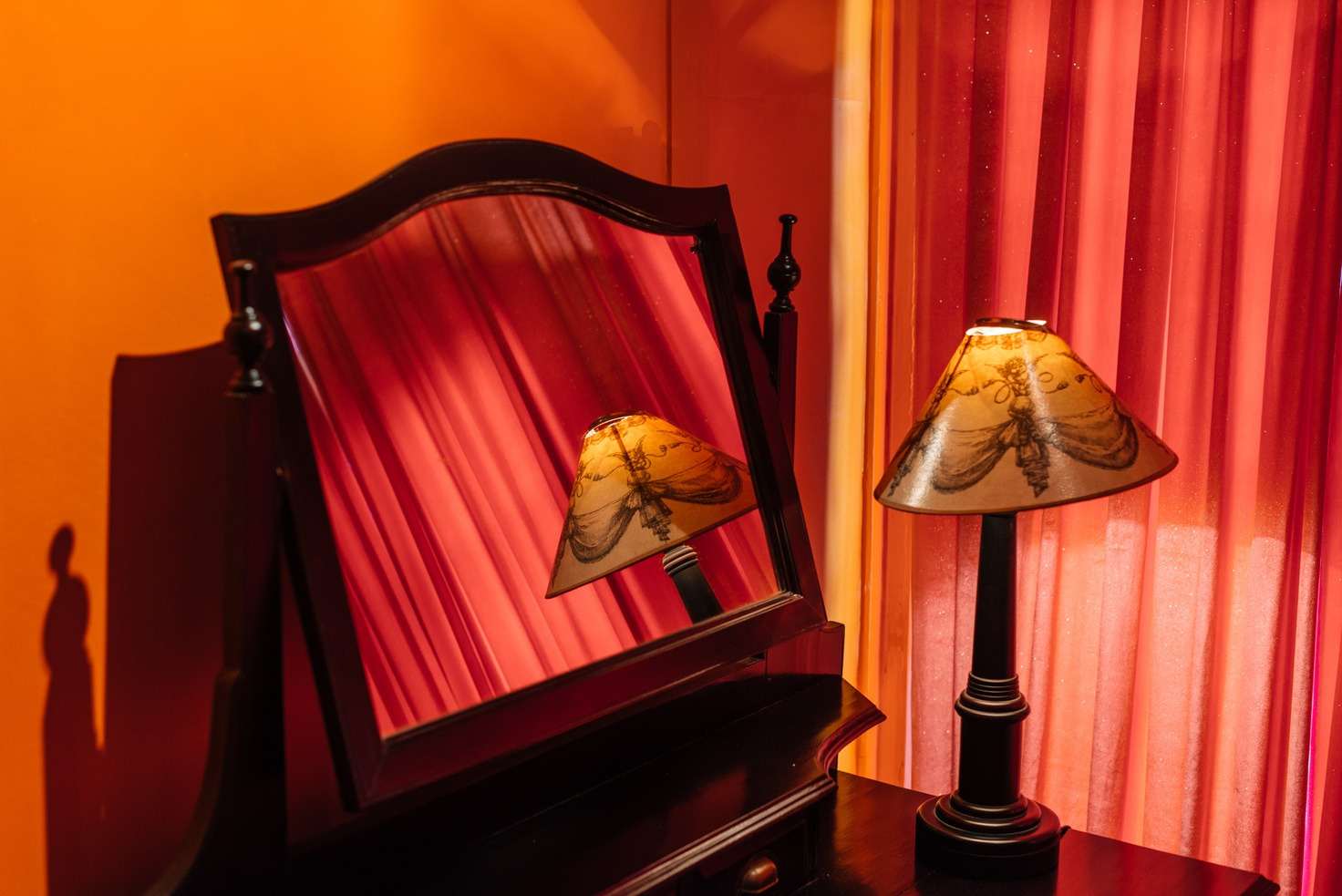 A dressing-table mirror reflecting red curtains and a moth-print lampshade.