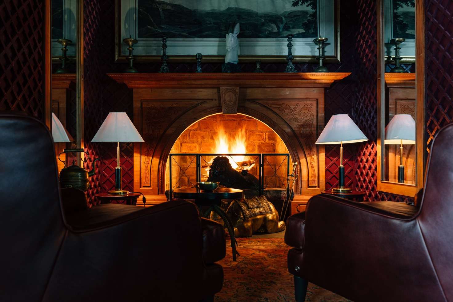 A dark snug with quilted oxblood walls, a carved timber fireplace with a fire burning, two armchairs and a brass camel lying at the hearth.