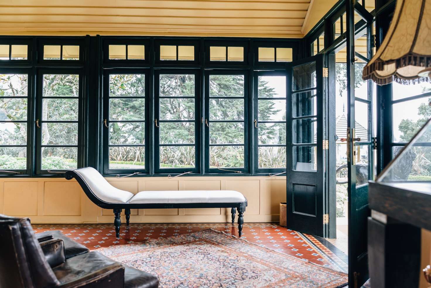 A glazed sunroom with green-black window frames, a white chaise longue and a patterned rug on a tiled floor.